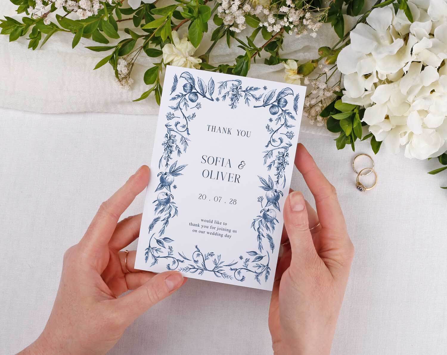 Person holding Blue Garden Wedding Thank You Cards surrounded by greenery and floral decorations.