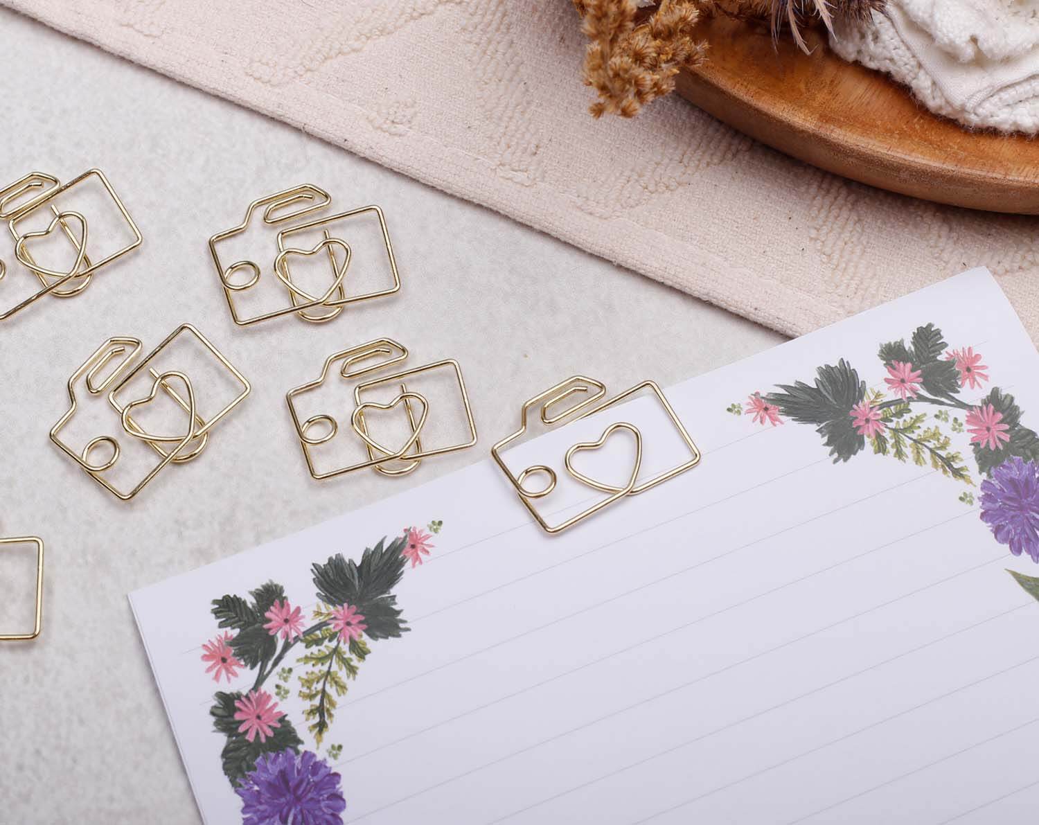 Gold Camera Paper Clips arranged on a notepad with floral design, showcasing their playful camera shape and creativity.