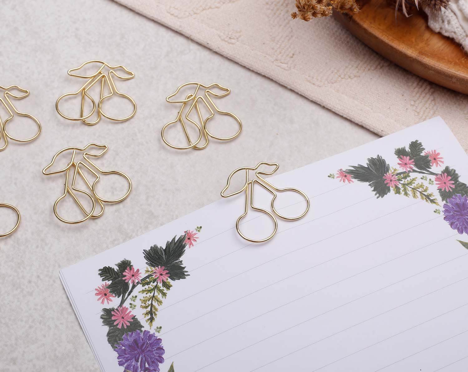 Gold Cherry Paper Clips arranged beside a floral notepad on a textured surface.