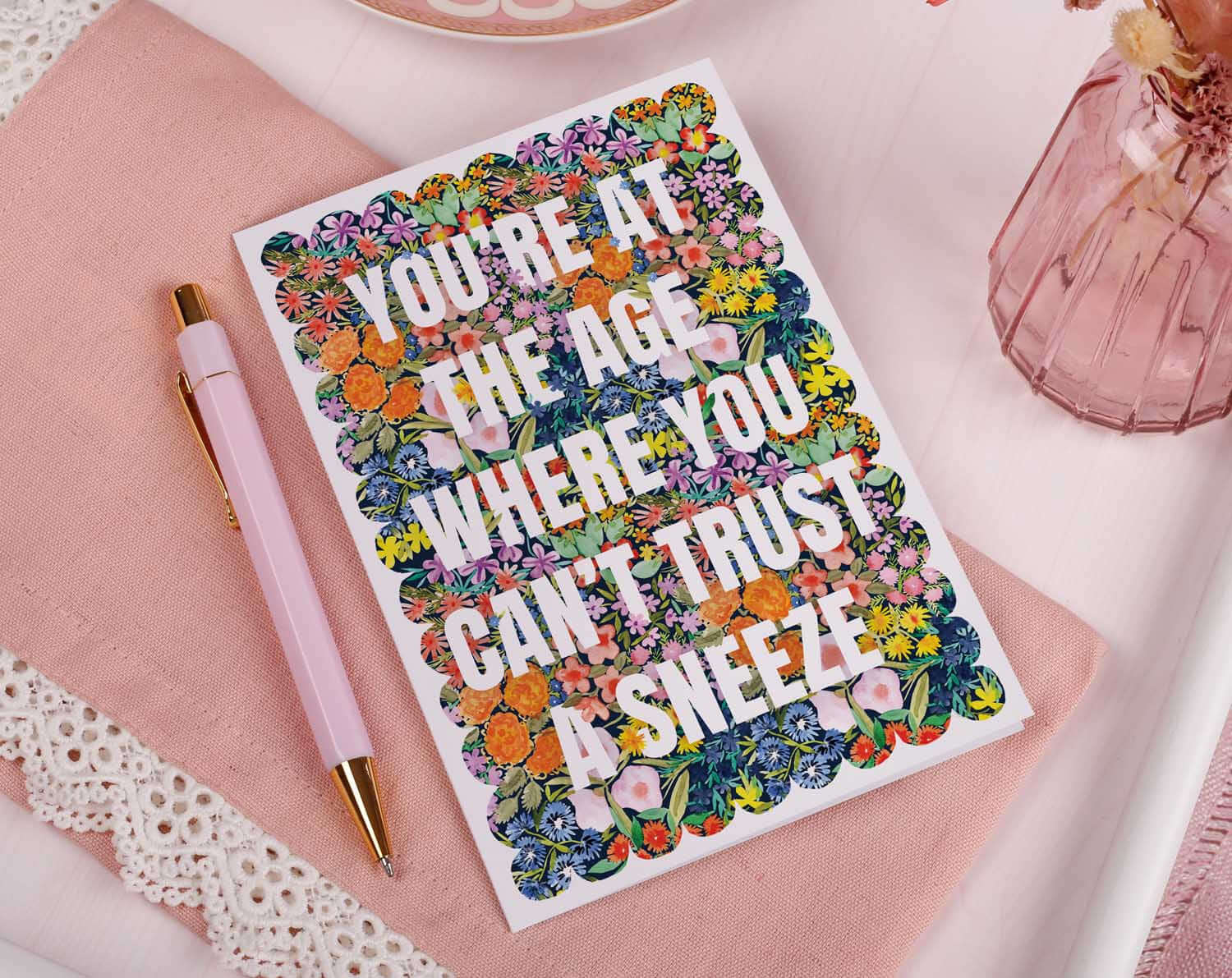 Joke Birthday Card with colorful floral background and joke text 'Can't Trust A Sneeze' on a table.