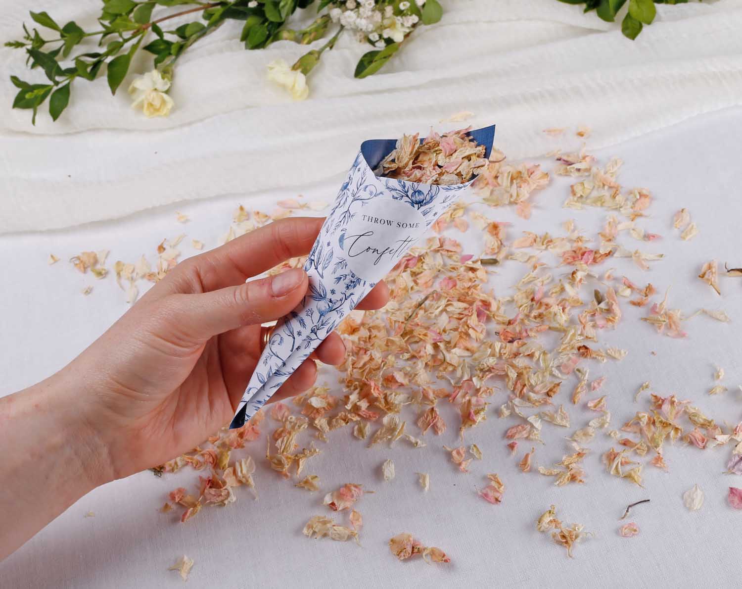 A hand holding Wedding Confetti Stickers cone filled with dried flower petals against a backdrop of scattered confetti.
