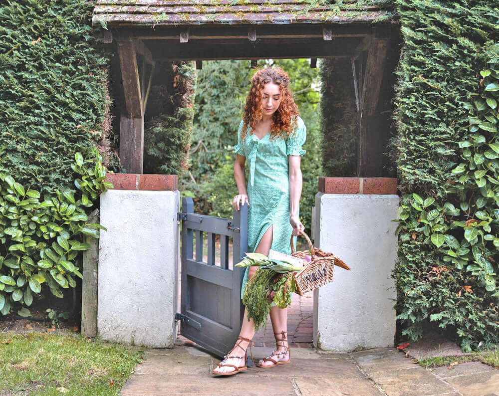 Woman in green dress standing at garden gate with basket of herbs, reflecting sunny stationery essentials.