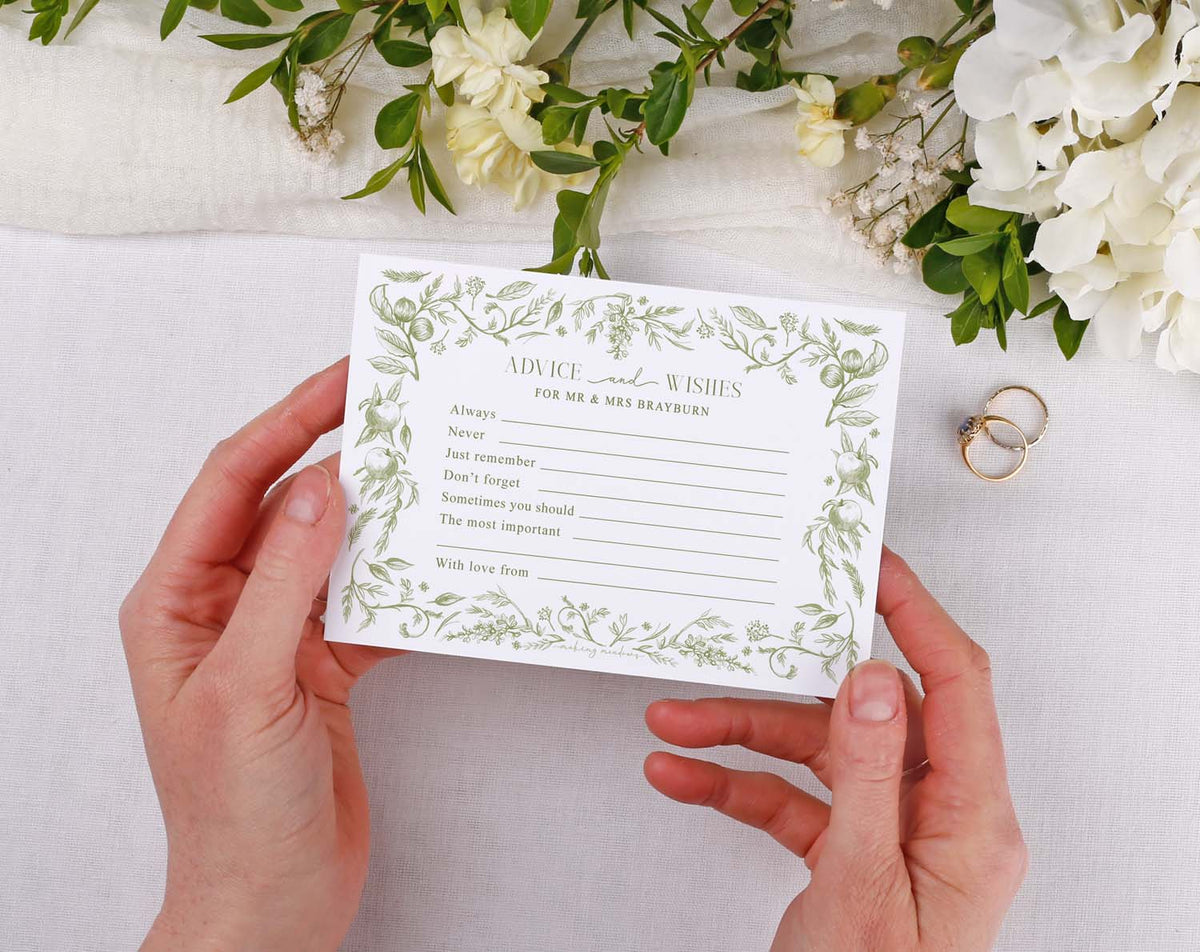 Person holding Sage Garden Wedding Advice Cards with floral design and rings on a table.