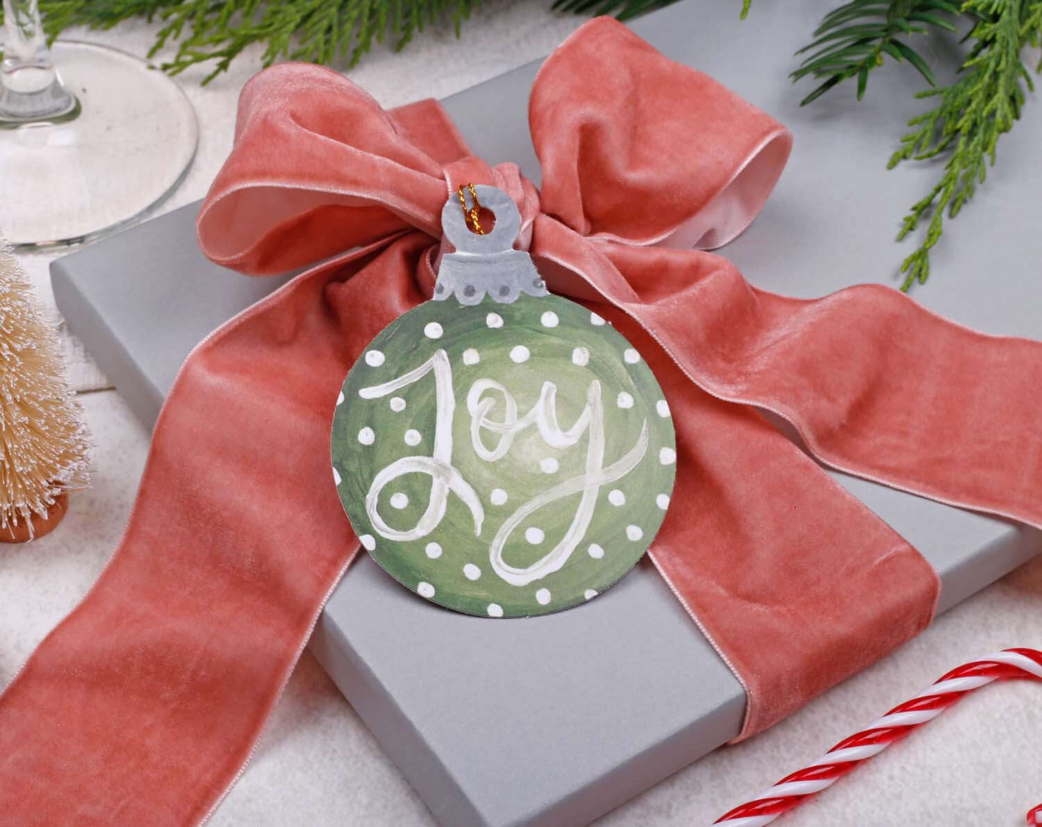 Bauble Christmas Gift Tags attached to a wrapped gift with a pink ribbon, featuring a green ornament saying 'Joy'.