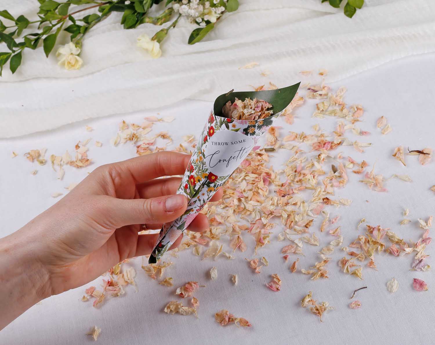 Hand holding Boho Wedding Confetti Cones filled with petals, surrounded by confetti on white table.