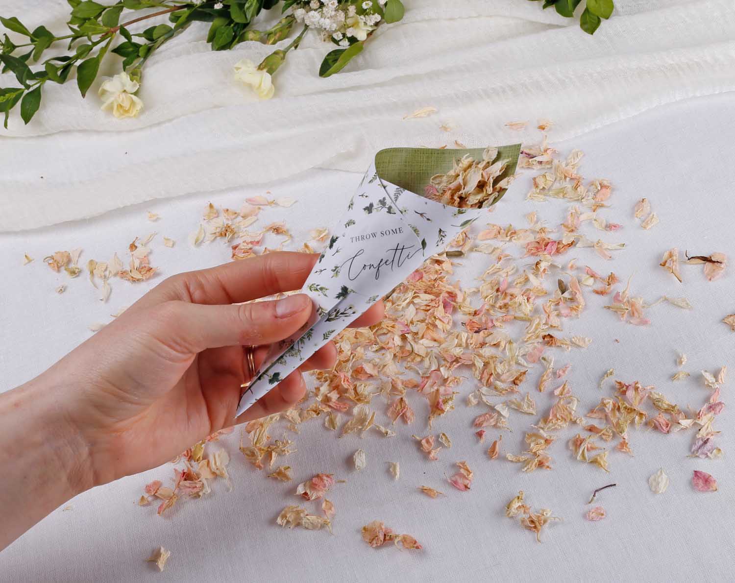 Botanical Wedding Confetti Cones held in hand with petals scattered on white fabric.