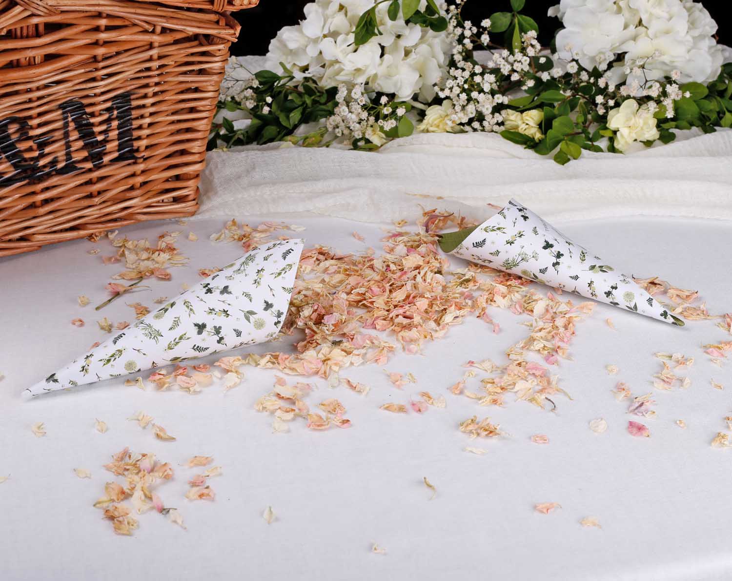 Botanical Wedding Confetti Cones displayed with floral confetti on a table by a wicker basket for a wedding ceremony.