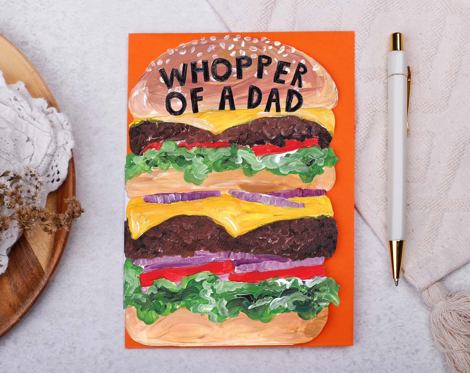 Card with a burger illustration and 'Whopper of a Dad' text on an orange background, placed on a textured surface with a pen and yarn.