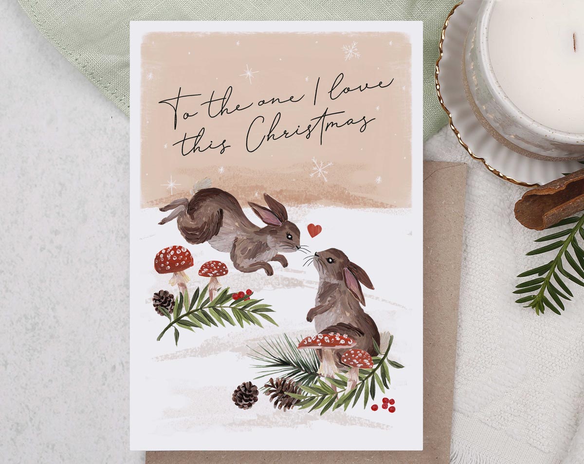 Christmas card, illustrated with a pair of loved up rabbits and a snow filled forest with foliage and mushrooms