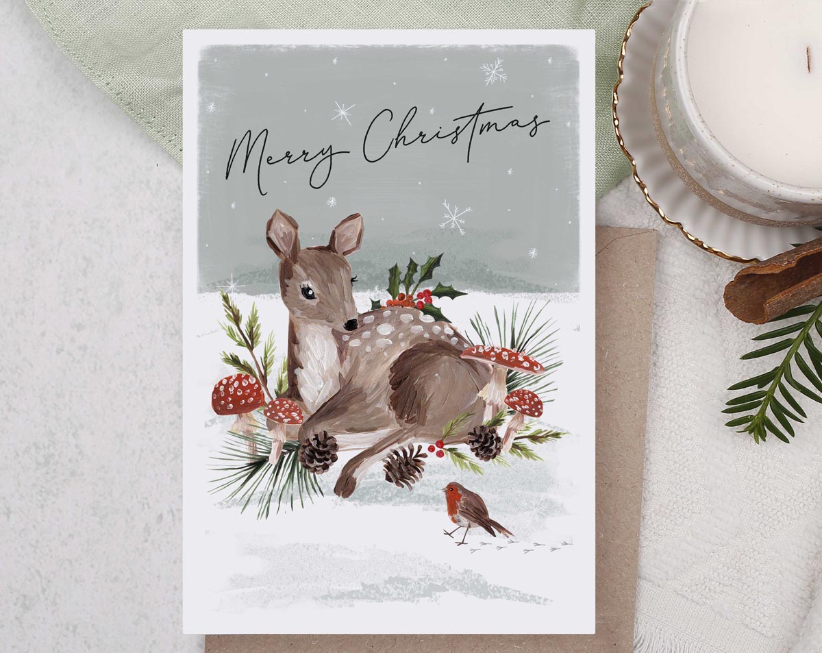 Christmas card, illustrated with a festive deer and robin, snow filled forest with foliage and mushrooms galore