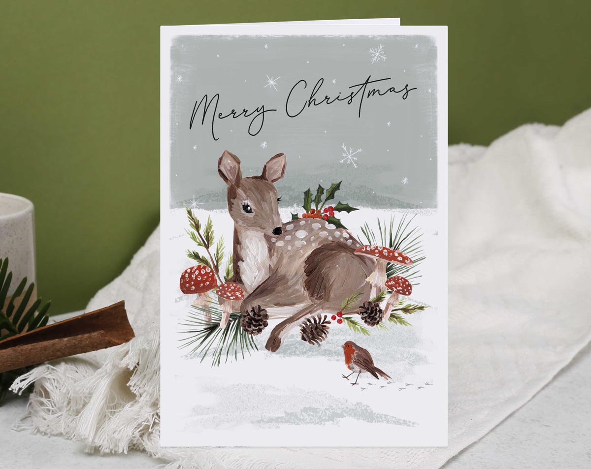Christmas card, illustrated with a festive deer and robin, snow filled forest with foliage and mushrooms galore