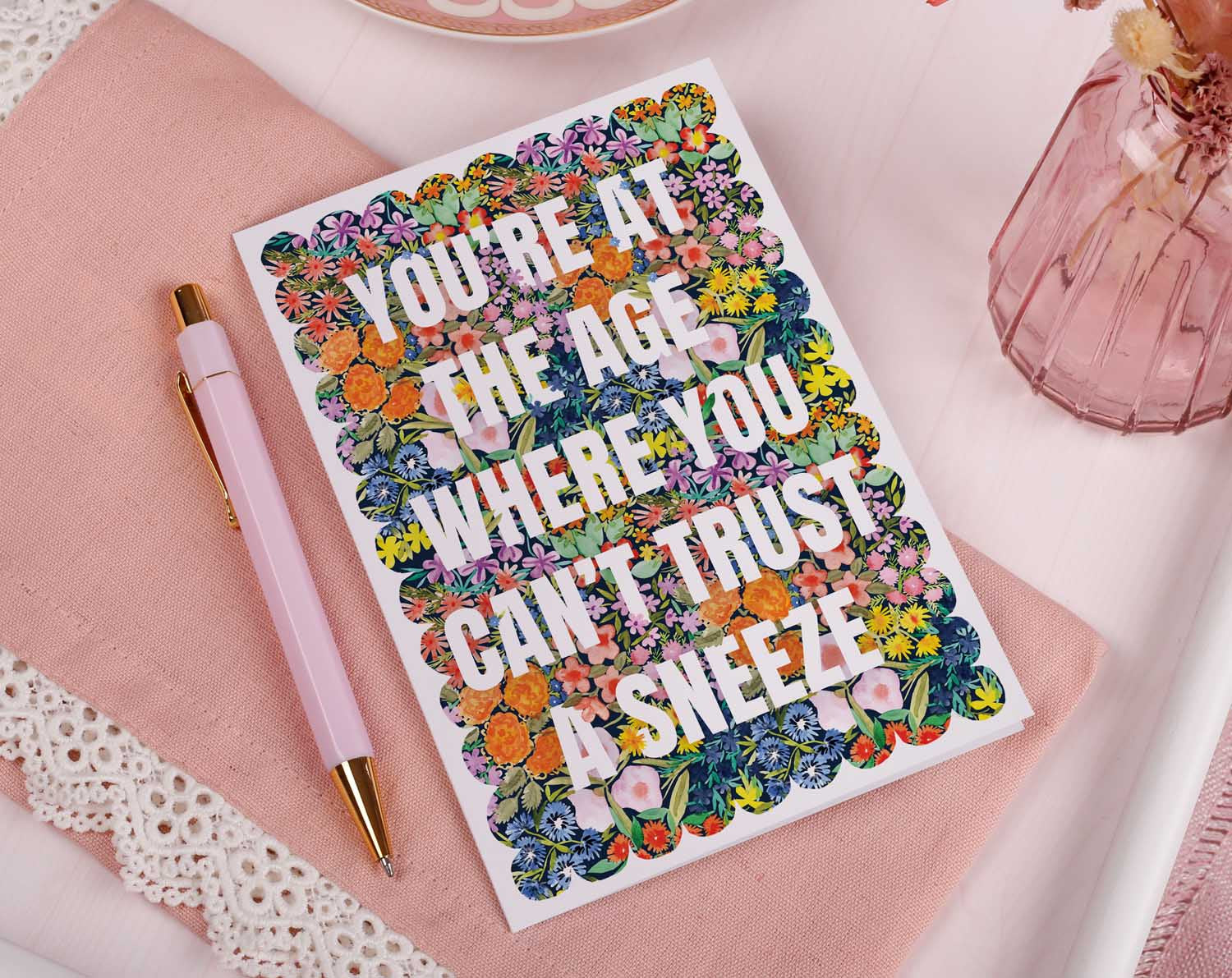 Joke Birthday Card with colorful floral background and joke text 'Can't Trust A Sneeze' on a table.