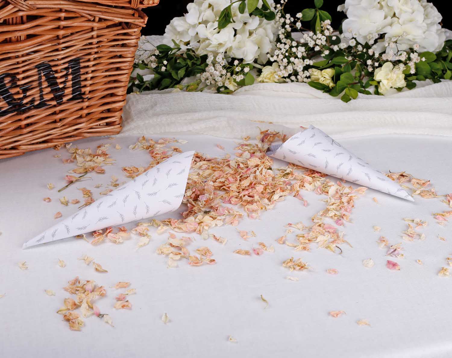 Two Minimalist Wedding Confetti Cones surrounded by flower petals on a table with a woven basket in the background.