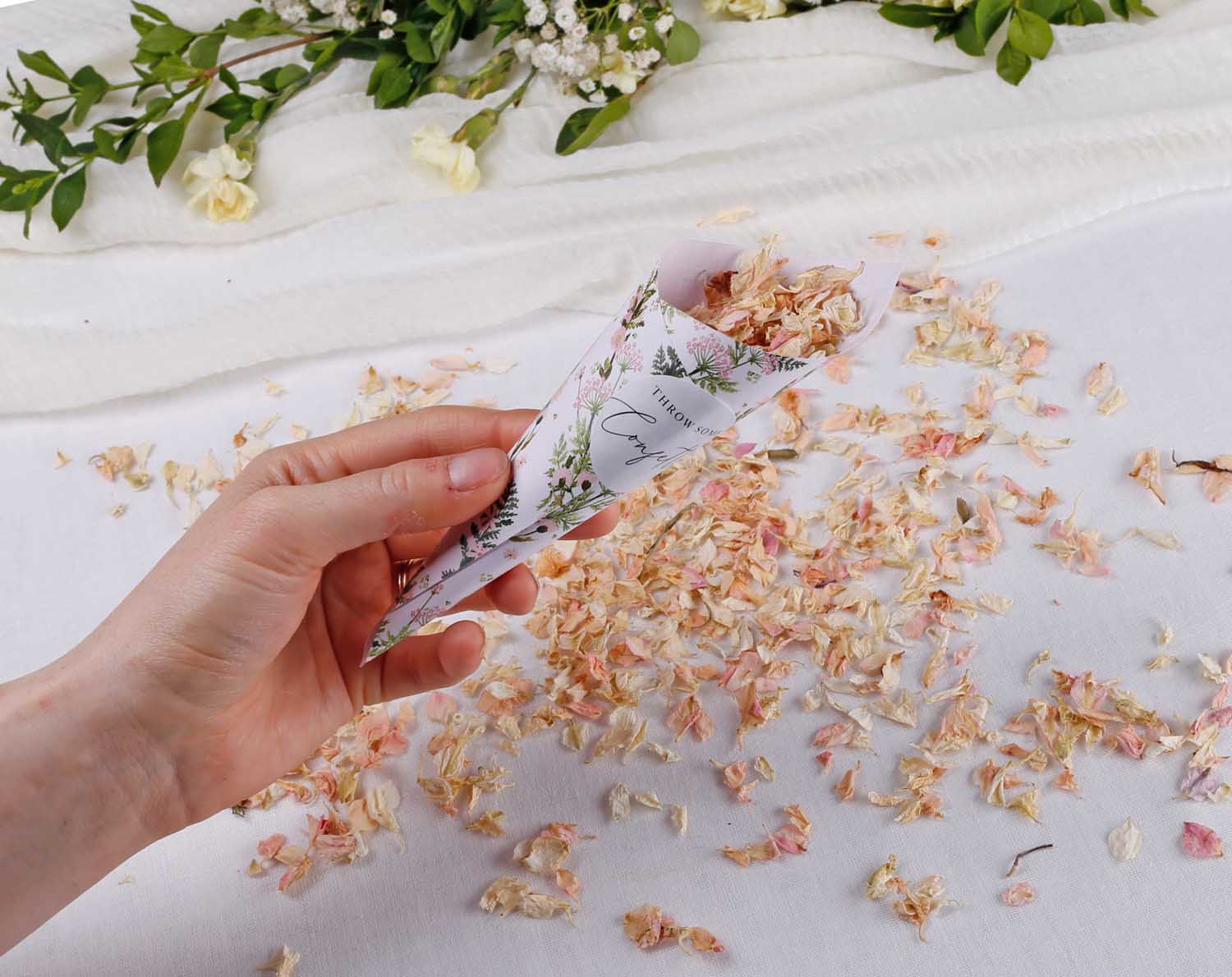 Pink Floral Wedding Confetti Cones filled with floral petals, held in a hand over scattered confetti on a white surface.
