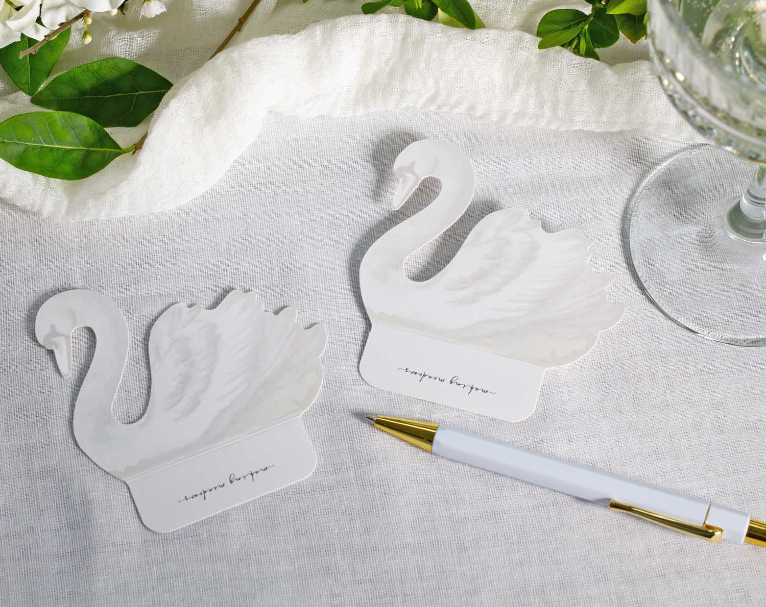 Swan Wedding Place Cards elegantly displayed on a table with green foliage and a pen.