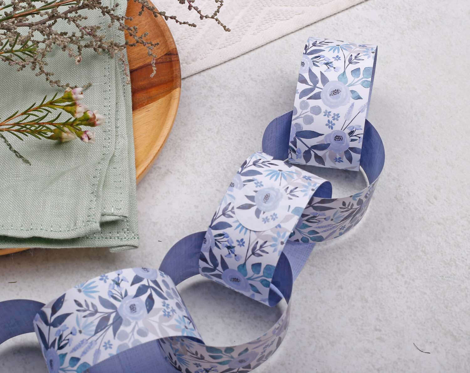 Blue Floral Paper Chains Garland displayed on a table with greenery and a wooden plate in the background.