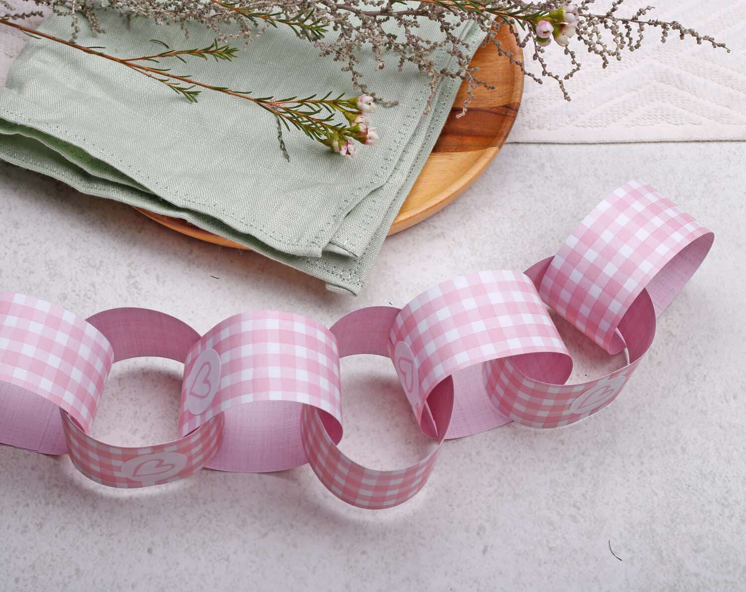 Pink Gingham Paper Chains Garland displayed on a rustic table with decorative green napkin and flowers.