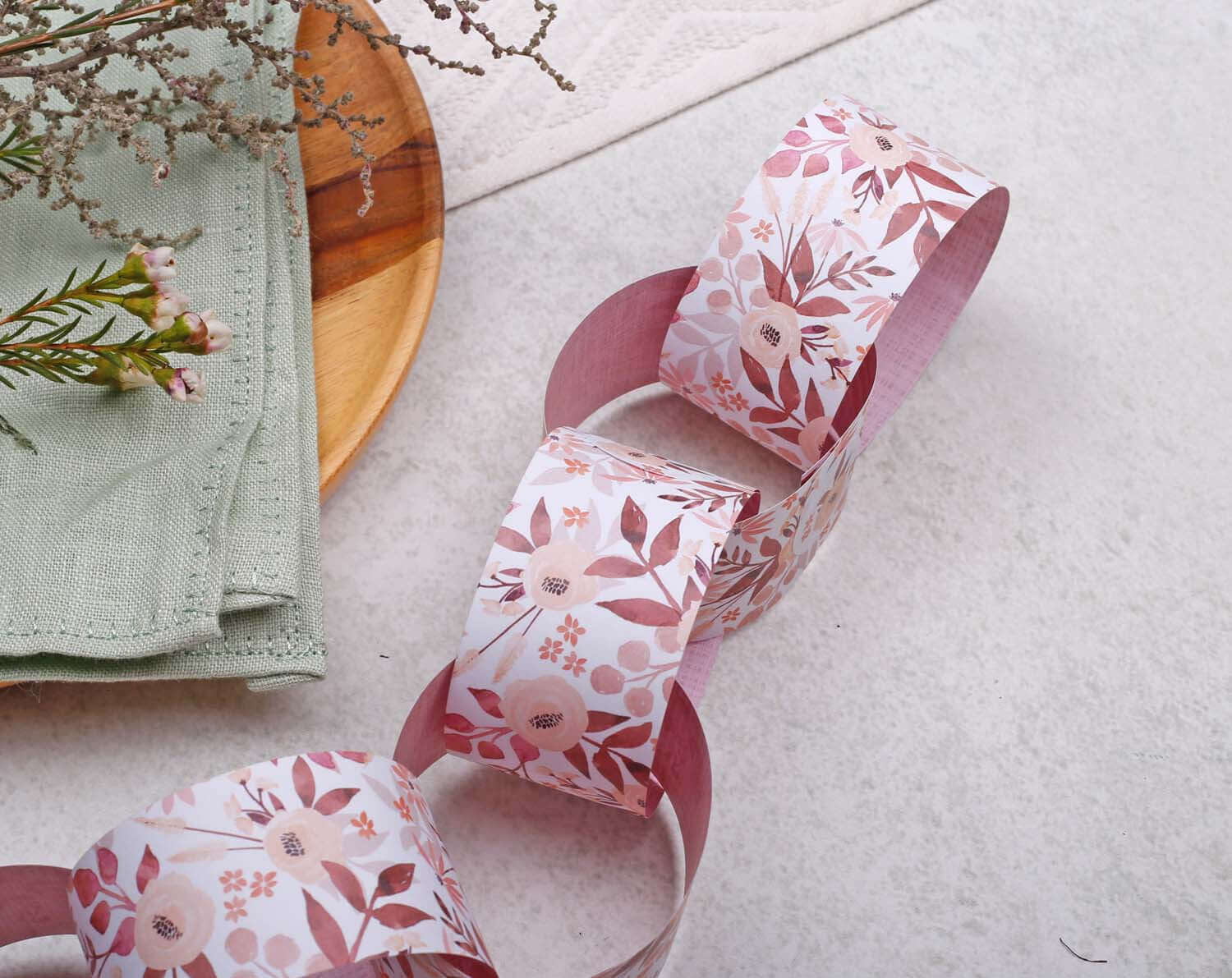 Pink Floral Paper Chains Garland displayed with a wooden plate and greenery on a light surface.