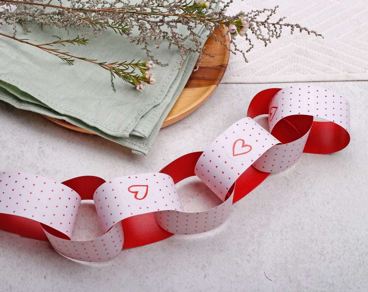 Pink Heart Paper Chains garland displayed on a wooden plate with green napkins and dried flowers.
