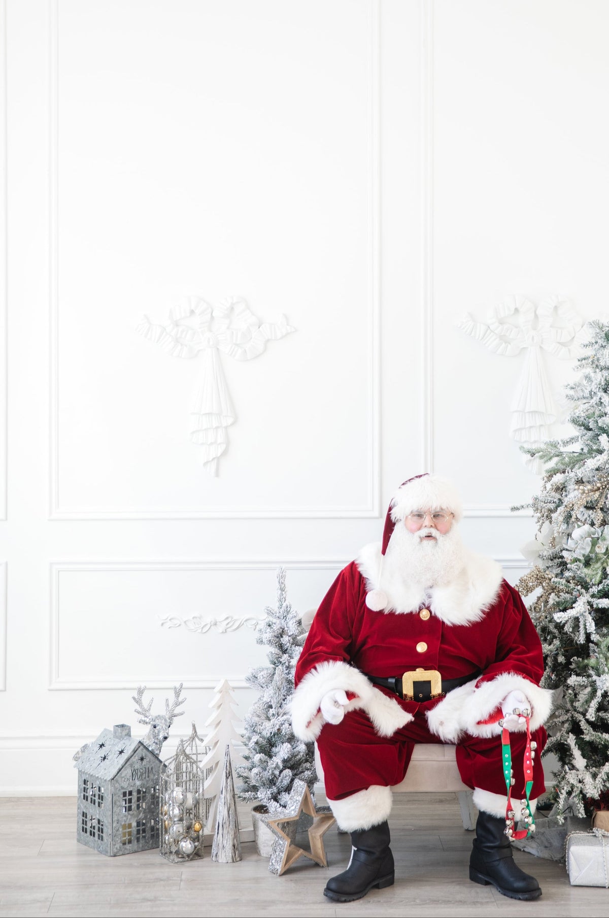 A man dressed as Santa Claus sits on a bench in a decorated holiday room. He wears a red velvet suit with white fur trim, black boots, and a gold belt, holding a candy cane.