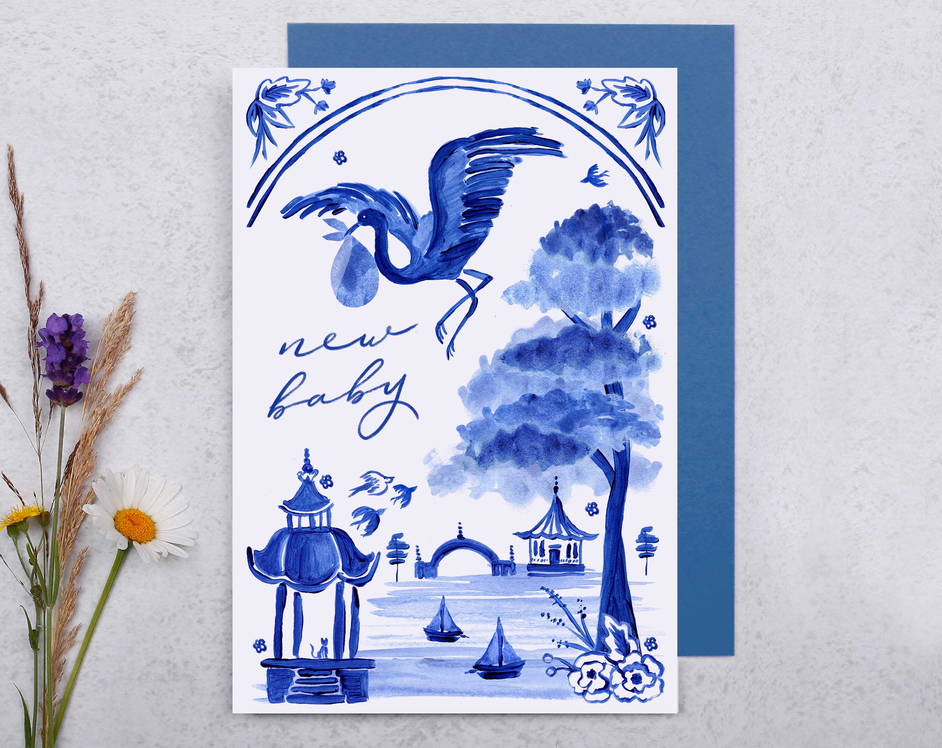 A blue porcelain inspired New Baby Card with a stork flying through the skies, over a beautiful landscape, while holding a baby in a blanket. 