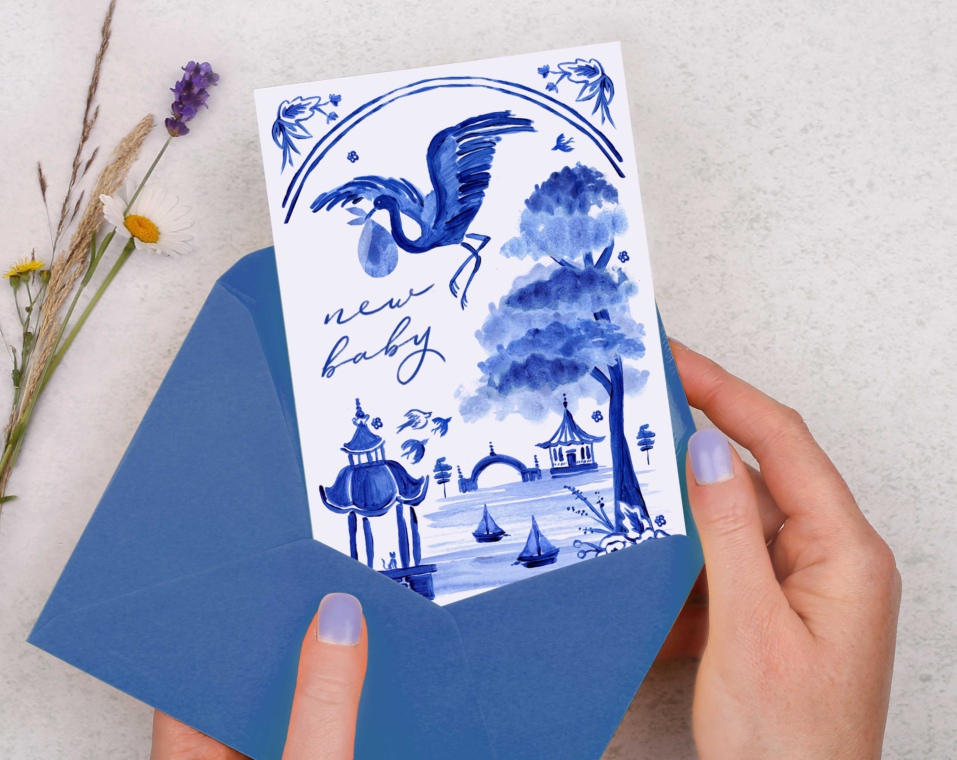 A blue porcelain inspired New Baby Card with a stork flying through the skies, over a beautiful landscape, while holding a baby in a blanket. 